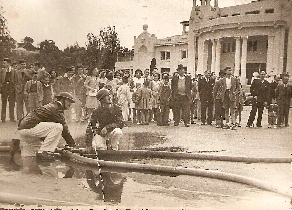 Fotografía sepia: dos bomberos con casco agachados junto a una manguera; detrás, una multitud observa frente a un edificio de columnas.